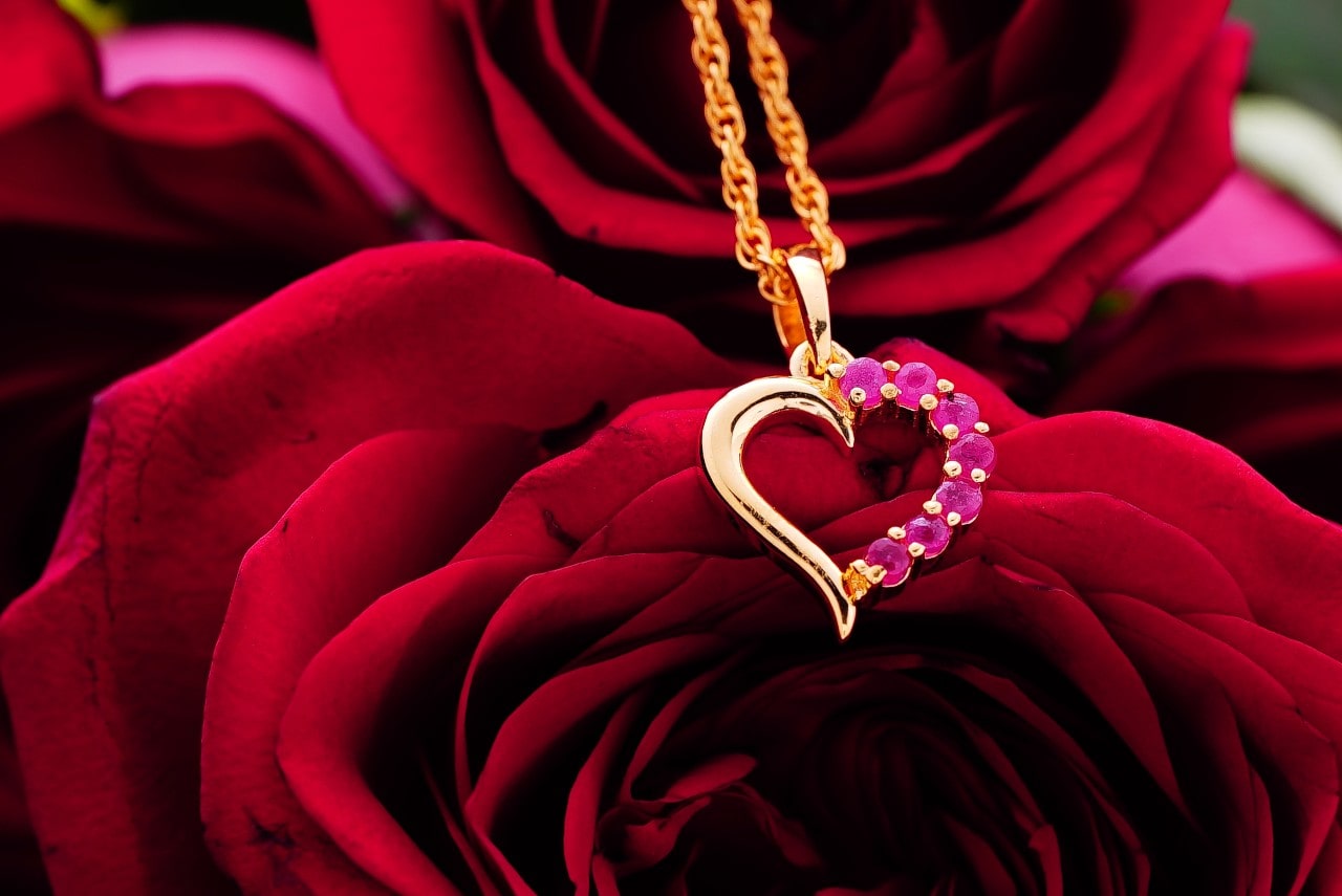 A yellow gold heart pendant with rubies along one side sitting on top of a bouquet of red roses