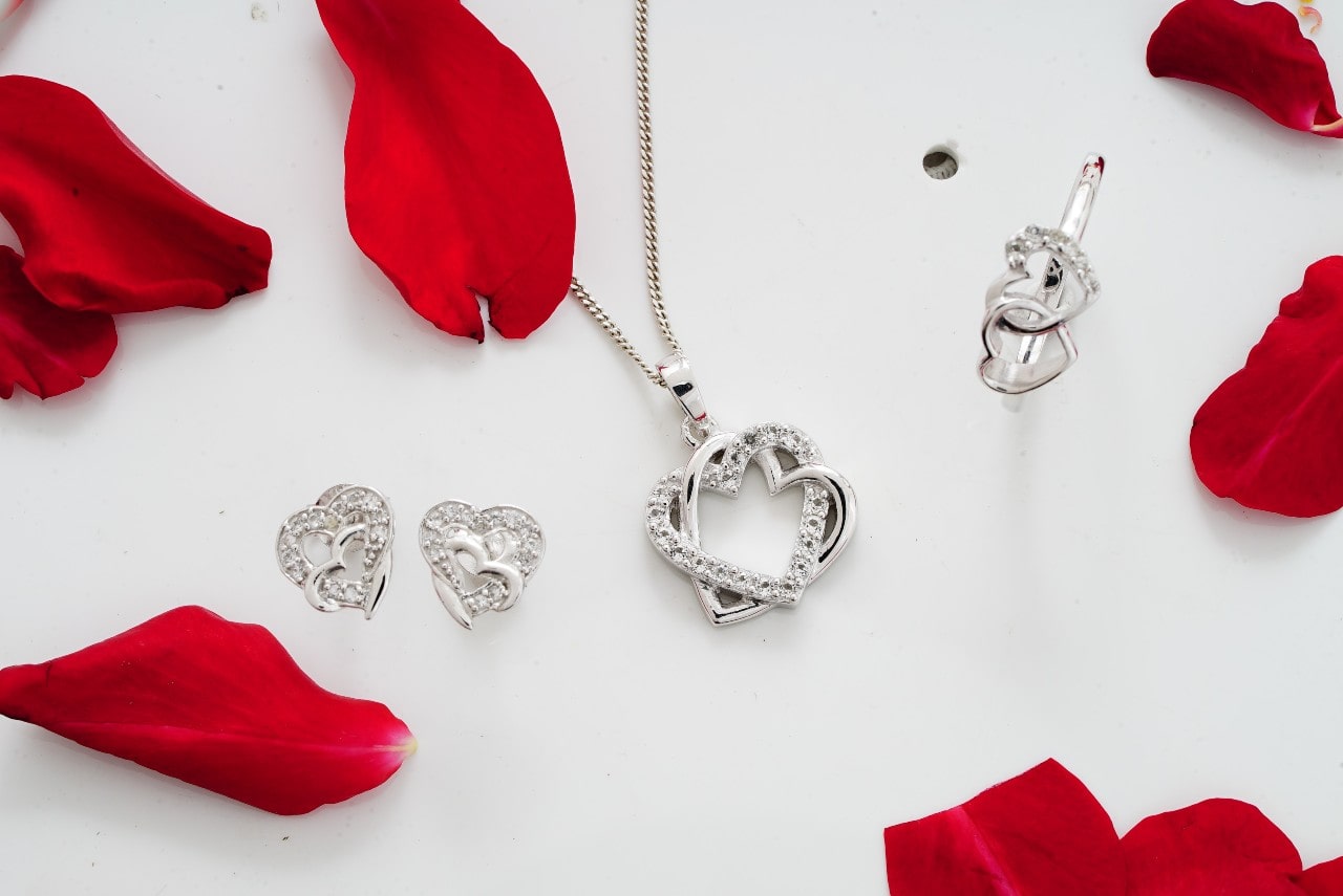 Double heart pendant, stud earrings, and fashion ring all detailed with diamonds and surrounded by red rose petals