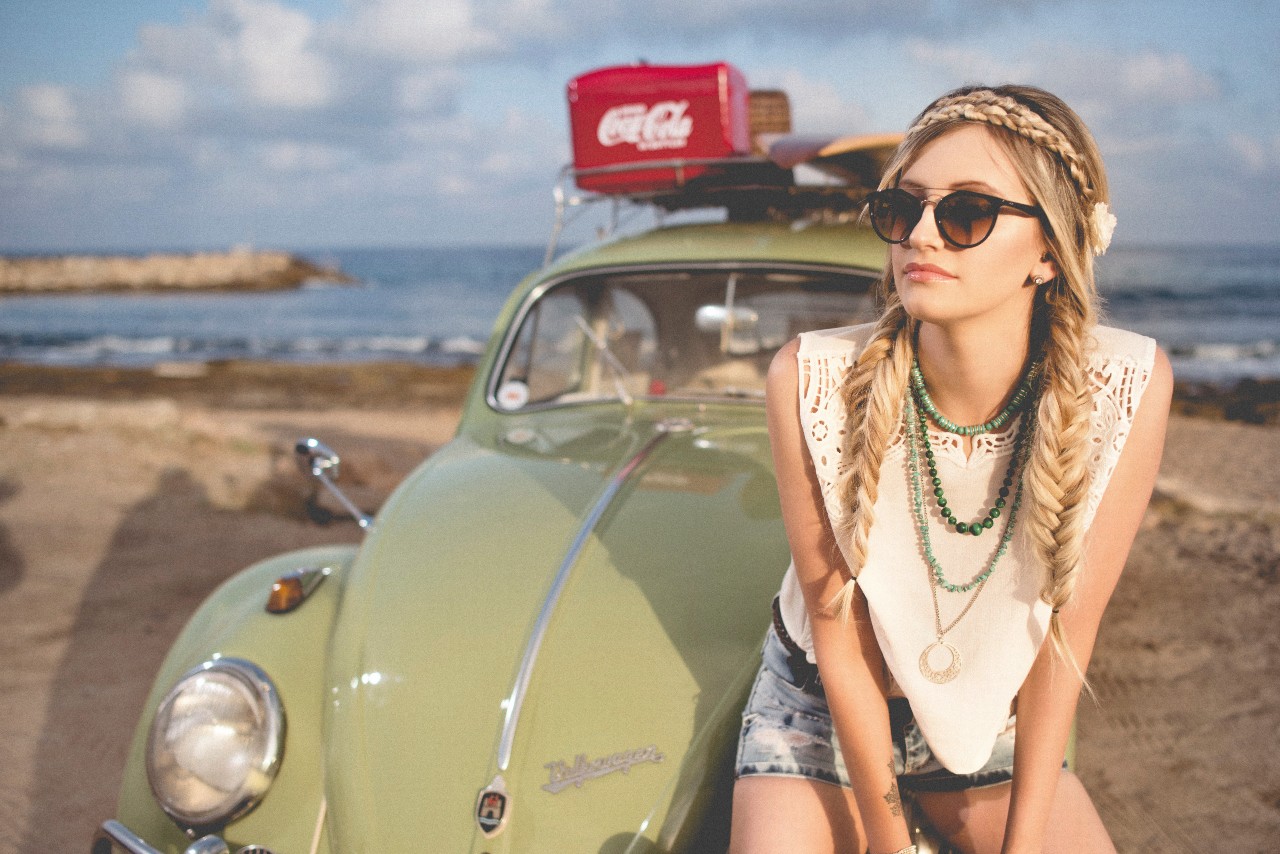 A woman sitting on the hood of her car on the beach, wearing multiple beaded and chain necklaces with a variety of textures, colours, and shapes