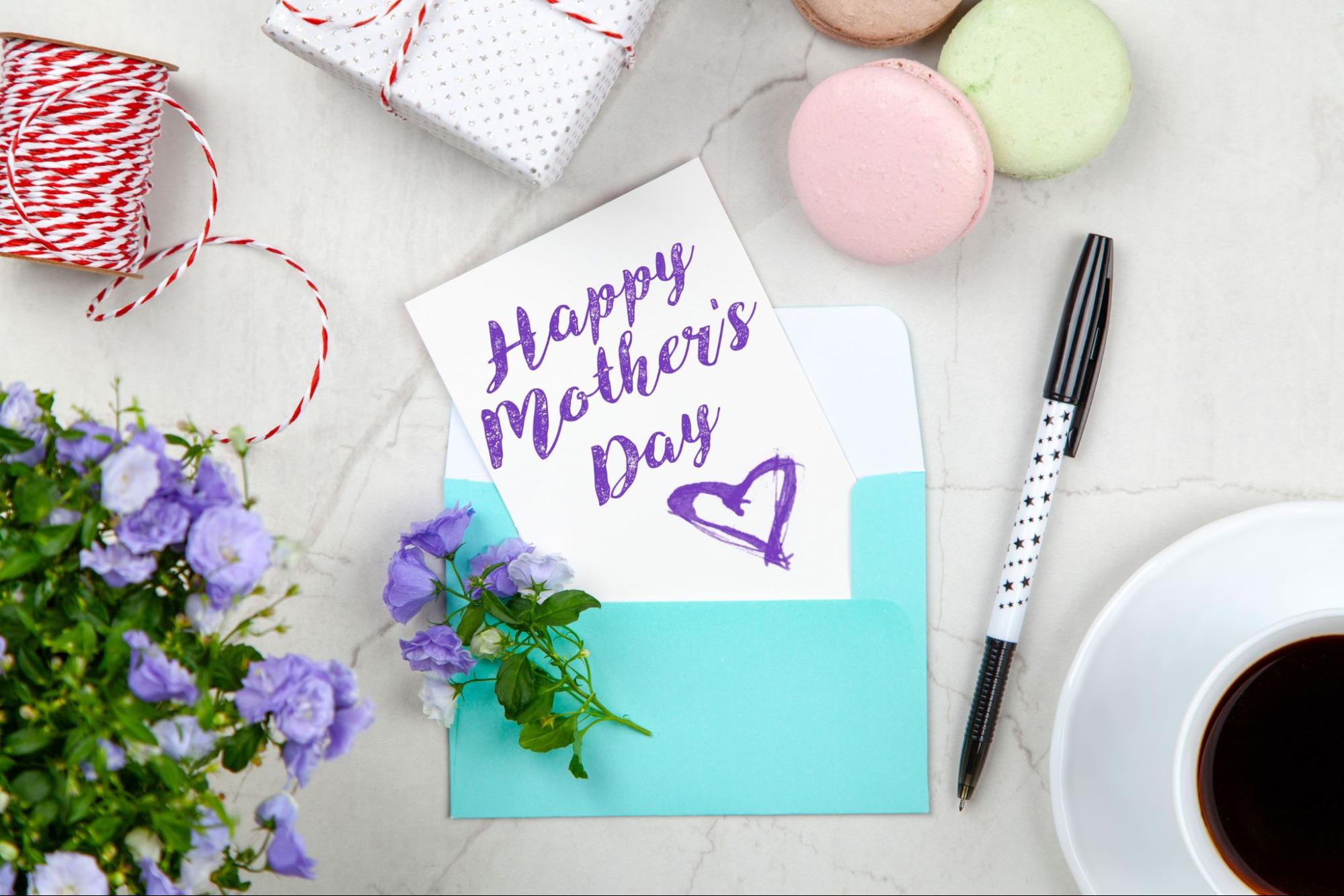 A handmade Mother?s Day card sits on a marble table with flowers, coffee, wrapped gifts, and macaroons