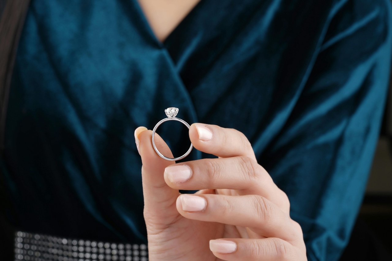 A woman wearing a velvet teal is holding a delicate diamond solitaire engagement ring.