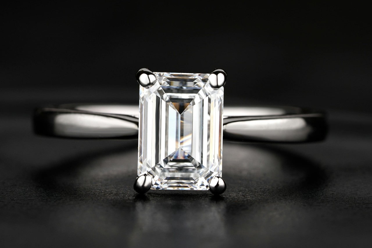 A close-up of an emerald-cut diamond engagement ring with a sleek silver band on a dark surface.