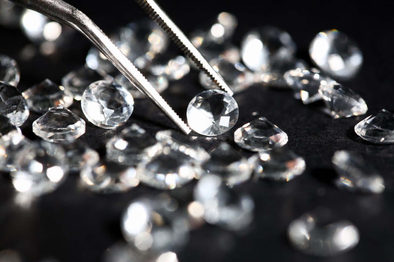 A close-up of several round cut diamonds on a soft black surface, with one being picked up with a pair of jeweler’s tweezers.