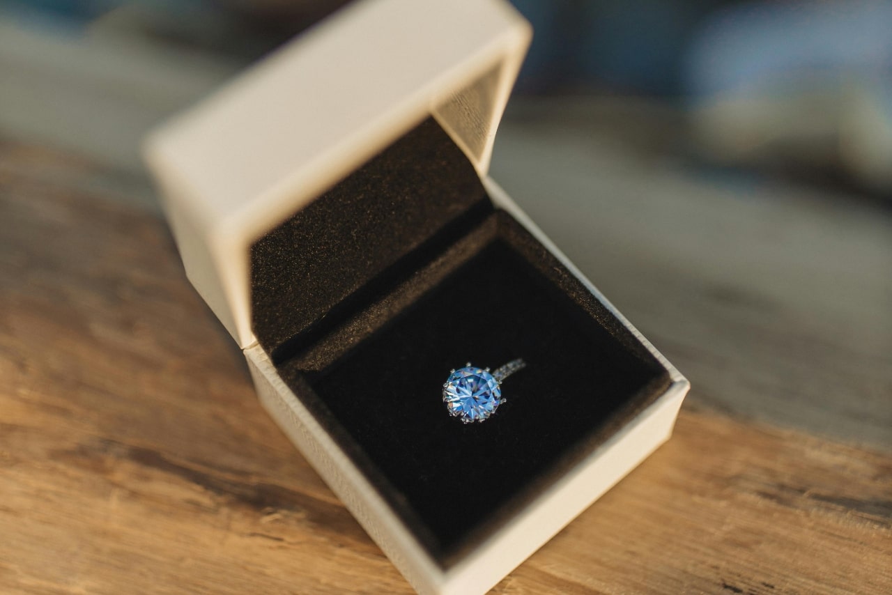 A close-up of an engagement ring with a blue centre stone in a white ring box. A close-up of an engagement ring with a blue centre stone in a white ring box.