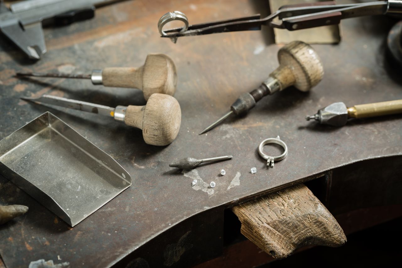 A jeweller's table full of tools, a white gold engagement ring setting, and several diamonds scattered nearby. A jeweller's table full of tools, a white gold engagement ring setting, and several diamonds scattered nearby.
